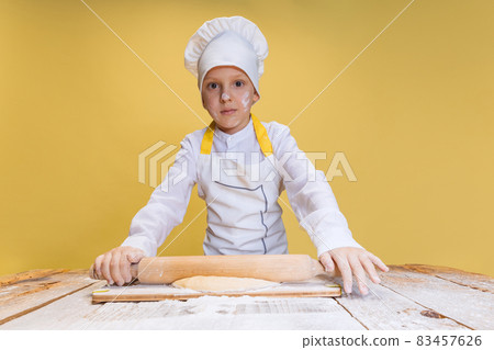 One cute boy wearing in white cook, chef jacket and hat kneads dough isolated on yellow studio background. One cute boy wearing in white cook, chef jacket and hat kneads dough isolated on yellow studio background. 83457626