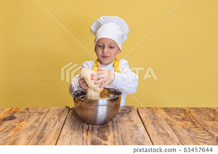 Cute smiling boy in white cook, chef jacket and hat kneads dough isolated on yellow studio background. 83457644