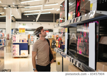 Male customer in casual clothes examining modern TV on shelf while shopping in spacious mall 83457904