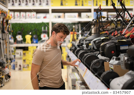 Side view of pensive casual male checking row of modern lawn mowers on shelf in power tools shop Side view of pensive casual male checking row of modern lawn mowers on shelf in power tools shop 83457907