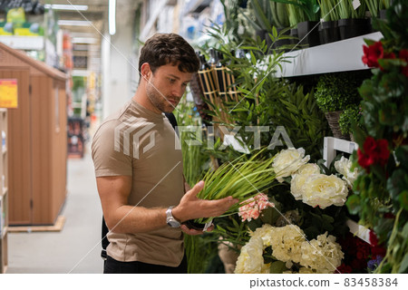 Male client in beige t shirt examining potted grass plant while standing near shelf with flowers in shop Male client in beige t shirt examining potted grass plant while standing near shelf with flowers in shop 83458384