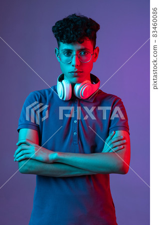 Cropped portrait of young boy wearing stylish glasses with headphones isolated over purple background in neon lights. Online education 83460086