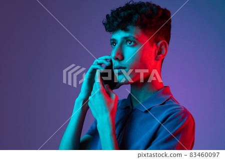 Cropped portrait of young boy in T-shirt talking on phone isolated over purple background in neon lights Cropped portrait of young boy in T-shirt talking on phone isolated over purple background in neon lights 83460097