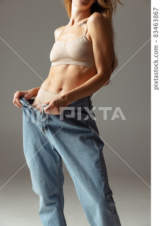 Portrait of young beautiful slim woman in blue jeans jumpersuit posing isolated over gray studio background. Weight Loss concept. Portrait of young beautiful slim woman in blue jeans jumpersuit posing isolated over gray studio background. Weight Loss concept. 83463867