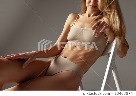 Cropped shot of young beautiful pretty woman in light underwear sitting on high chair isolated over gray studio background. Natural beauty concept. 83463874