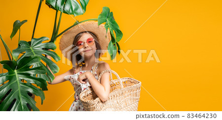Pre-teen girl in straw hat, orange sunglasses holds straw handbag while stands near tropical plants Pre-teen girl in straw hat, orange sunglasses holds straw handbag while stands near tropical plants 83464230