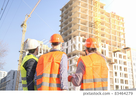 Male workers engineers at construction site in helmets hardhats look at high-rise building skyscraper. Engineers are discussing the progress of the construction of commercial real estate. Male workers engineers at construction site in helmets hardhats look at high-rise building skyscraper. Engineers are discussing the progress of the construction of commercial real estate. 83464279
