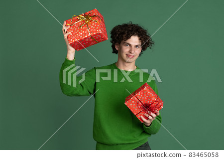 Cropped portrait of young man in green sweater with two gift boxes isolated over dark green background. New Year celebration Cropped portrait of young man in green sweater with two gift boxes isolated over dark green background. New Year celebration 83465058