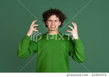 Cropped portrait of young man in green sweater with angry, irritating facial expression isolated over dark green background 83465074