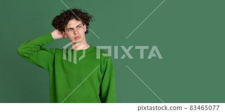 Cropped portrait of young man in green sweater with facial expression of misunderstanding isolated over dark green background Cropped portrait of young man in green sweater with facial expression of misunderstanding isolated over dark green background 83465077