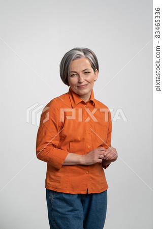 Happy middle aged woman looking at camera in orange shirt and denim pants. Pretty mid aged grey haired woman in orange shirt isolated on grey background Happy middle aged woman looking at camera in orange shirt and denim pants. Pretty mid aged grey haired woman in orange shirt isolated on grey background 83465336