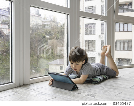 Curious boy watch cartoons on digital tablet. Kid lies on floor and uses electronic device. Indoor leisure for children while it's raining outside. 83465487