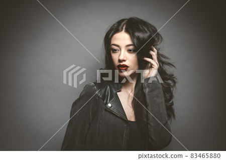 Fashion woman with beautiful face in a leather jacket with beautiful long dark hair isolated on gray background. Young female model with professional make-up. Cosmetics Fashion woman with beautiful face in a leather jacket with beautiful long dark hair isolated on gray background. Young female model with professional make-up. Cosmetics 83465880