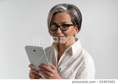 Portrait of a grey haired mature woman in white shirt and eye glasses using digital tablet. The use of technology by the elderly browsing internet shopping online on pad isolated on white background Portrait of a grey haired mature woman in white shirt and eye glasses using digital tablet. The use of technology by the elderly browsing internet shopping online on pad isolated on white background 83465881