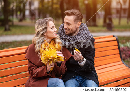 Embracing man found a small leaf happy couple sitting on the bench hugged in park wearing coats and scarfs collecting a bouquet of fallen leaves. Love story concept. Tinted image 83466202