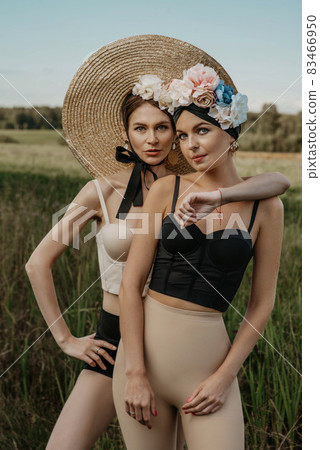 Girls in tops and shorts, in a hat and with a rim of flowers, posing against the background of nature. The first girl holds the stems of the reed. Another girl with a basket in which different flowers Girls in tops and shorts, in a hat and with a rim of flowers, posing against the background of nature. The first girl holds the stems of the reed. Another girl with a basket in which different flowers 83466950