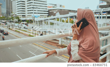 A young Muslim girl in a pink hijab talking on the phone above road traffic in the city center. 83468731