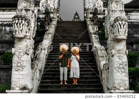 A couple stands on the stairs of the Baltic temple and cover their faces with rice caps. Man and woman traveling in Indonesia. Couple at the Bali gate. The couple travels the world. Tourists in Bali A couple stands on the stairs of the Baltic temple and cover their faces with rice caps. Man and woman traveling in Indonesia. Couple at the Bali gate. The couple travels the world. Tourists in Bali 83469252