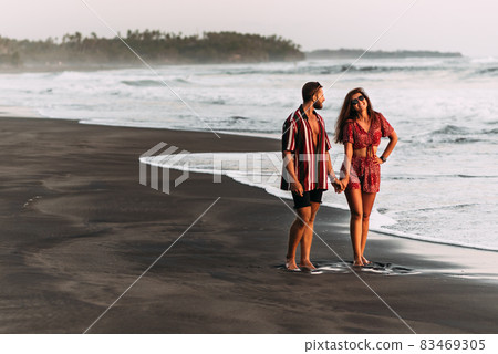 Happy couple holding hands walking on the sandy beach. Couple in love at sunset by the sea. Couple in love on vacation. Honeymoon trip. Romantic couple enjoying a beach walk at sunset 83469305