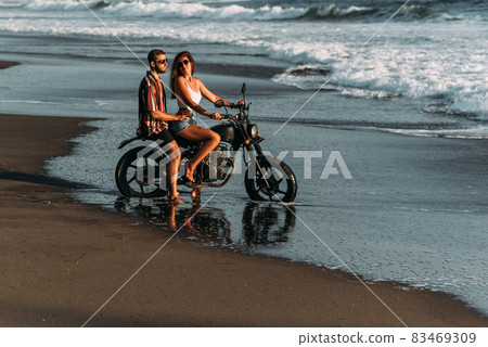 Couple on a motorcycle on the beach. A couple in love on the beach meets the sunset. A man embraces a woman on the beach. Beautiful couple on a motorcycle. Travel on a motorcycle. Lovers on the beach Couple on a motorcycle on the beach. A couple in love on the beach meets the sunset. A man embraces a woman on the beach. Beautiful couple on a motorcycle. Travel on a motorcycle. Lovers on the beach 83469309