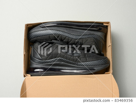pair of black textile sneakers in an open brown paper box on a gray table, top view 83469356