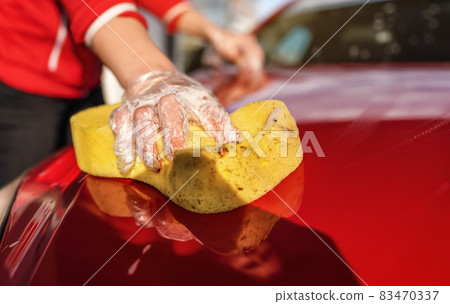 Young woman cleaning front hood of her car, closeup detail on yellow sponge held by hand in plastic glove 83470337