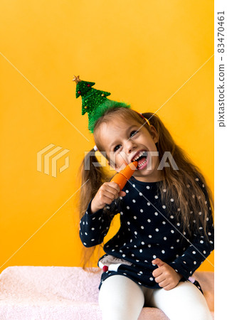 Portrait positive cheerful smiling happy little schoolgirl girl Christmas tree decoration polka dot dress biting eat orange carrots on orange background. New year, holiday, celebration, winter concept Portrait positive cheerful smiling happy little schoolgirl girl Christmas tree decoration polka dot dress biting eat orange carrots on orange background. New year, holiday, celebration, winter concept 83470461