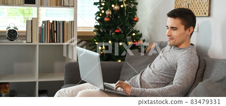 Smiling man sitting on couch near Christmas tree and using laptop computer. Smiling man sitting on couch near Christmas tree and using laptop computer. 83479331