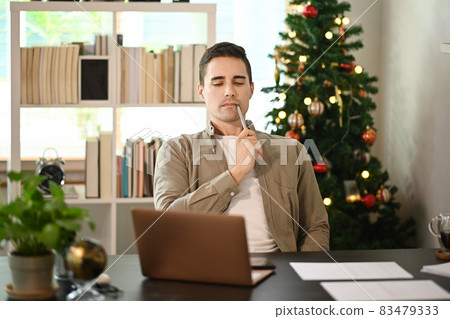 Young man sitting near Christmas tree and using laptop computer. Young man sitting near Christmas tree and using laptop computer. 83479333