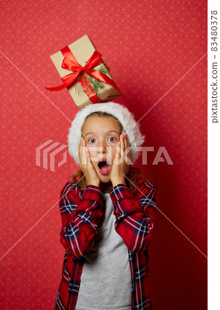 Cute little girl in santa hat with a Christmas gift on her head 83480378