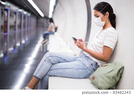 Woman in mask with smartphone sitting on bench in subway station Woman in mask with smartphone sitting on bench in subway station 83482243