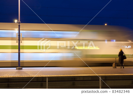 KOUVOLA, FINLAND - NOVEMBER 8, 2018: Long exposure photo. Train in motion on the station at night. Blurred woman with travel bag staying on the platform. KOUVOLA, FINLAND - NOVEMBER 8, 2018: Long exposure photo. Train in motion on the station at night. Blurred woman with travel bag staying on the platform. 83482346