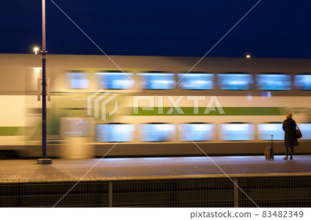 KOUVOLA, FINLAND - NOVEMBER 8, 2018: Long exposure photo. Train in motion on the station at night. Blurred woman with travel bag staying on the platform. KOUVOLA, FINLAND - NOVEMBER 8, 2018: Long exposure photo. Train in motion on the station at night. Blurred woman with travel bag staying on the platform. 83482349