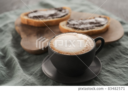 Cappuccino in black cup with ciabatta slices with chocolate spread on background Cappuccino in black cup with ciabatta slices with chocolate spread on background 83482571