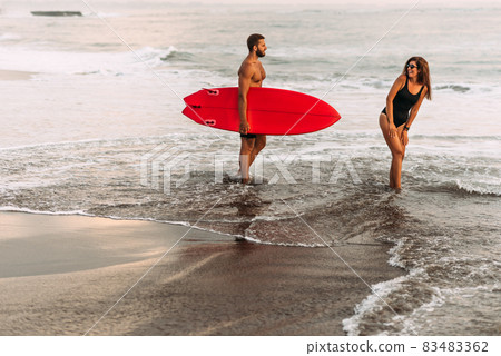 Beautiful couple have fun at the sea. Couple in love on the beach with a surfboard at sunset. Beautiful couple on the island of Bali. Couple of surfers walking on coast in Indonesia. Vacation at sea 83483362