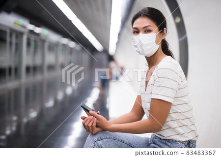 Woman in face mask with smartphone sitting on bench in subway station Woman in face mask with smartphone sitting on bench in subway station 83483367