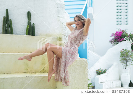 Beautiful caucasian young woman with blond hair sitting and holding her hair. Posing barefoot. Smiling with the teeth and looking at camera. 83487308