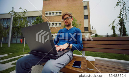 Working from park. Handsome student studying on computer at campus outdoors and drinks coffee. 83488524