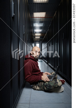 The hacker with a bald head and a beard sits with the computer among servers, the burglar, Storage of data, cloudy services, system breaking, attack of hackers The hacker with a bald head and a beard sits with the computer among servers, the burglar, Storage of data, cloudy services, system breaking, attack of hackers 83493097