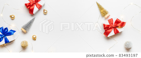 Christmas gift. White gift with red bow, gold balls and sparkling lights garland in xmas decoration on white background for greeting card. Xmas backdrop with space for text. Christmas gift. White gift with red bow, gold balls and sparkling lights garland in xmas decoration on white background for greeting card. Xmas backdrop with space for text. 83493197