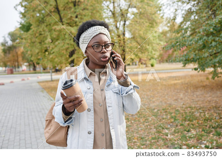 African young woman walking in the park during her coffee break and talking on mobile phone African young woman walking in the park during her coffee break and talking on mobile phone 83493750