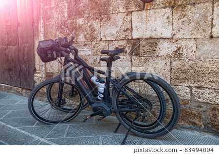 San Quirico d'Orcia, Tuscany, Italy. August 2020. Two ebikes parked on the pavement, one has a flat front wheel. San Quirico d'Orcia, Tuscany, Italy. August 2020. Two ebikes parked on the pavement, one has a flat front wheel. 83494004