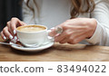 Side view of women hands with white cup of black coffe on light wooden table background. Media. Closeup shot of delicate girls hands taking small cup of coffee. 83494022