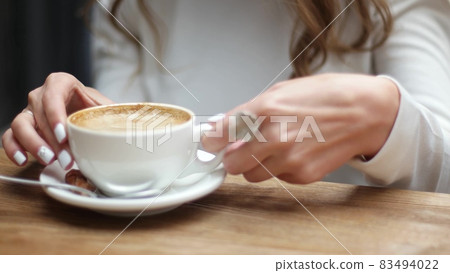 Side view of women hands with white cup of black coffe on light wooden table background. Media. Closeup shot of delicate girls hands taking small cup of coffee. Side view of women hands with white cup of black coffe on light wooden table background. Media. Closeup shot of delicate girls hands taking small cup of coffee. 83494022