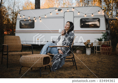 Beautiful caucasian woman is sitting wrapped in a plaid and drinking a warming drink outdoors. Traveling in a motor home in autumn. 83494361