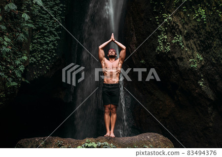 A man of athletic build does yoga. Healthy lifestyle. The concentration of the body. A man does yoga in nature. A man does yoga in Bali. A man meditates in nature. Meditation at the waterfall 83494376