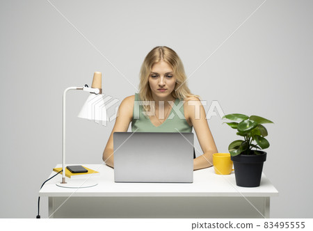 Portrait of a pretty young woman studying while sitting at the table with grey laptop computer, notebook. Smiling business woman working with a laptop isolated on a grey background. Portrait of a pretty young woman studying while sitting at the table with grey laptop computer, notebook. Smiling business woman working with a laptop isolated on a grey background. 83495555