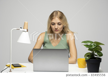 Smiling business woman working with a laptop isolated on a grey background. Portrait of a pretty young woman studying while sitting at the table with grey laptop computer, notebook. Smiling business woman working with a laptop isolated on a grey background. Portrait of a pretty young woman studying while sitting at the table with grey laptop computer, notebook. 83495556