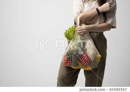 Zero waste concept. Young brunet woman holding reusable mesh cotton shopping bag with groceries from a market. Concept of no plastic. Zero waste, plastic free. Sustainable lifestyle. 83495567