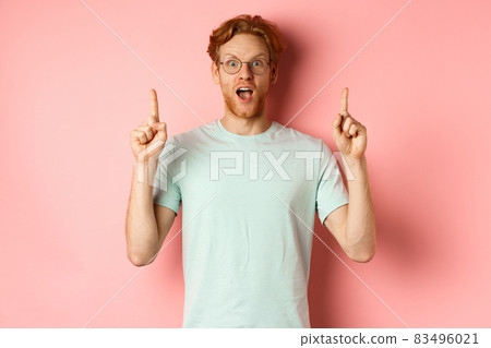 Portrait of surprised guy checking out advertisement, gasping amazed and pointing fingers up, showing promo, standing over pink background 83496021